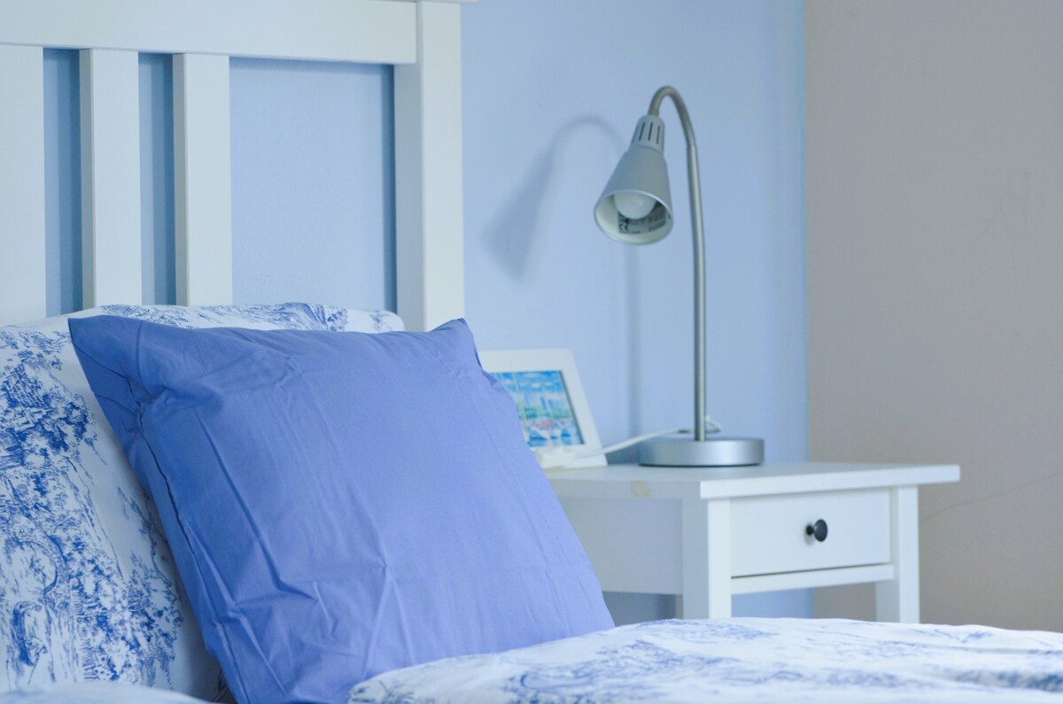 A spacious bed with a soft blue pillow is positioned against a light blue wall. A bedside table holds a sleek lamp and a digital photo frame, offering convenient lighting and decoration.