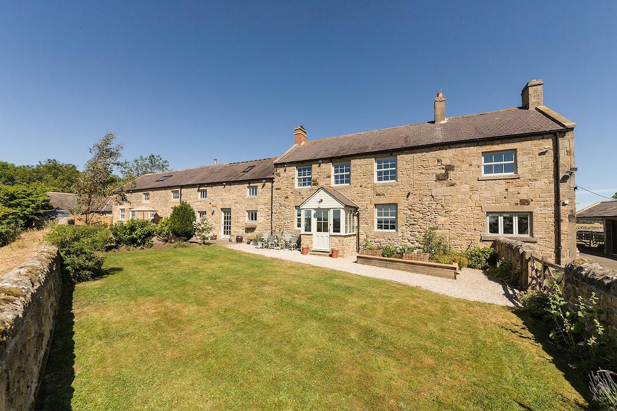 The exterior showcases a traditional stone farmhouse set within a spacious lawn, featuring a well-maintained garden area with bushes and seasonal plants. Sunlight bathes the building, highlighting large windows and a conservatory that invites natural light into the home.