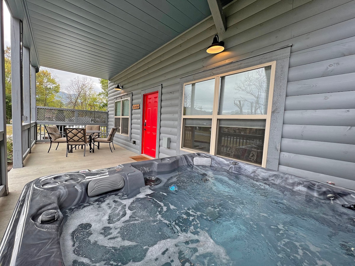 An outdoor hot tub is positioned on a covered deck, featuring relaxing bubbling water. In the background, a table and chairs offer an inviting space for gatherings. A bright red door adds a pop of color to the light gray log cabin exterior.