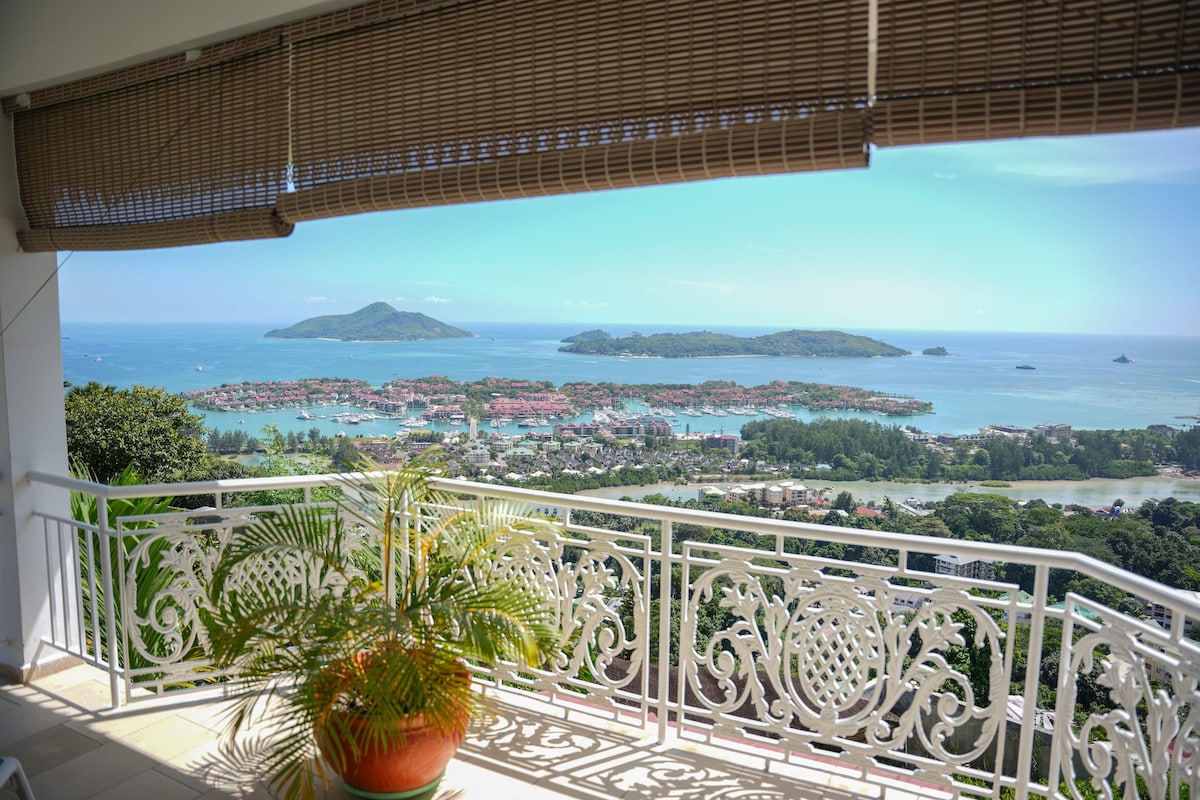 A spacious private terrace features a decorative railing and a large potted plant. Breathtaking views of the sea and nearby islands are visible in the distance, complemented by a clear blue sky and soft white clouds.