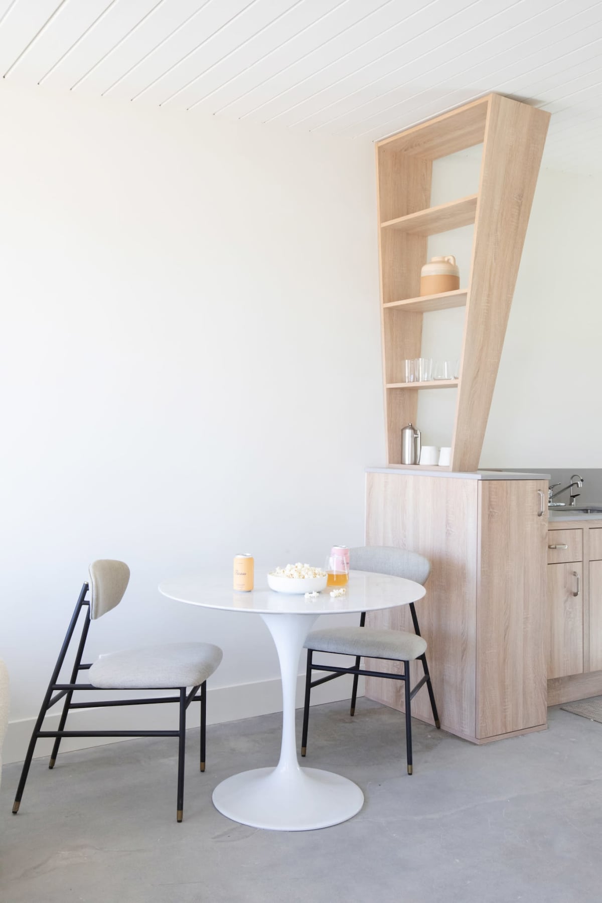 A small dining area is featured, showcasing a round white table surrounded by two chairs. A wood cabinetry unit with open shelving is visible in the background, housing simple dishware. The kitchenette area includes a countertop with a small sink and appliances, providing a functional living space.