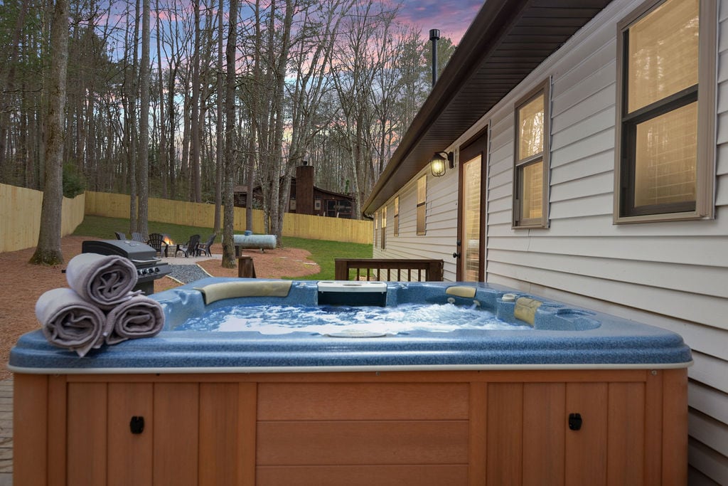 A hot tub is situated on a wooden deck, surrounded by nature and tall trees, offering a serene atmosphere. Rolled towels are neatly arranged beside the tub. The backyard features a fire pit in the distance, with additional seating visible.