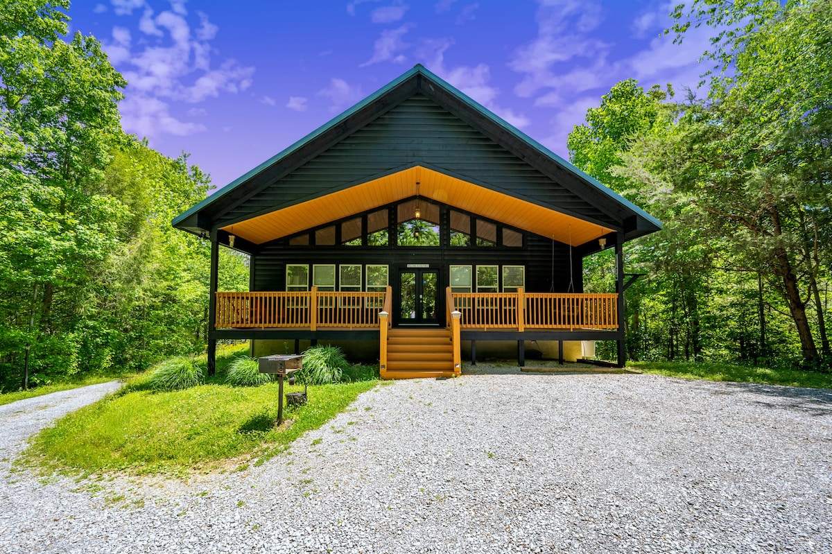 A charming cabin features a modern exterior with a combination of dark wood and striking orange accents. The spacious front porch is adorned with wooden railings and provides a welcoming entrance, surrounded by lush greenery and gravel pathways leading to the entrance.