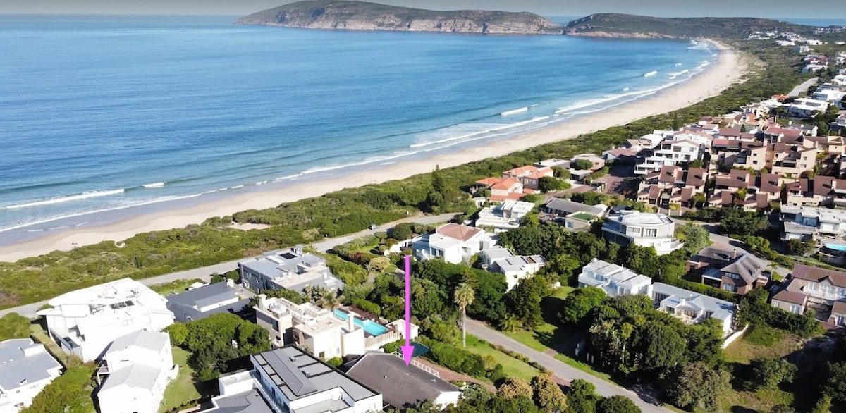 An aerial view captures the villa's location, showcasing proximity to Sanctuary Beach and the Robberg Peninsula. The expansive coastline meets lush greenery, with nearby residential areas visible. The serene ocean waves and sandy shore create a tranquil backdrop for the property.