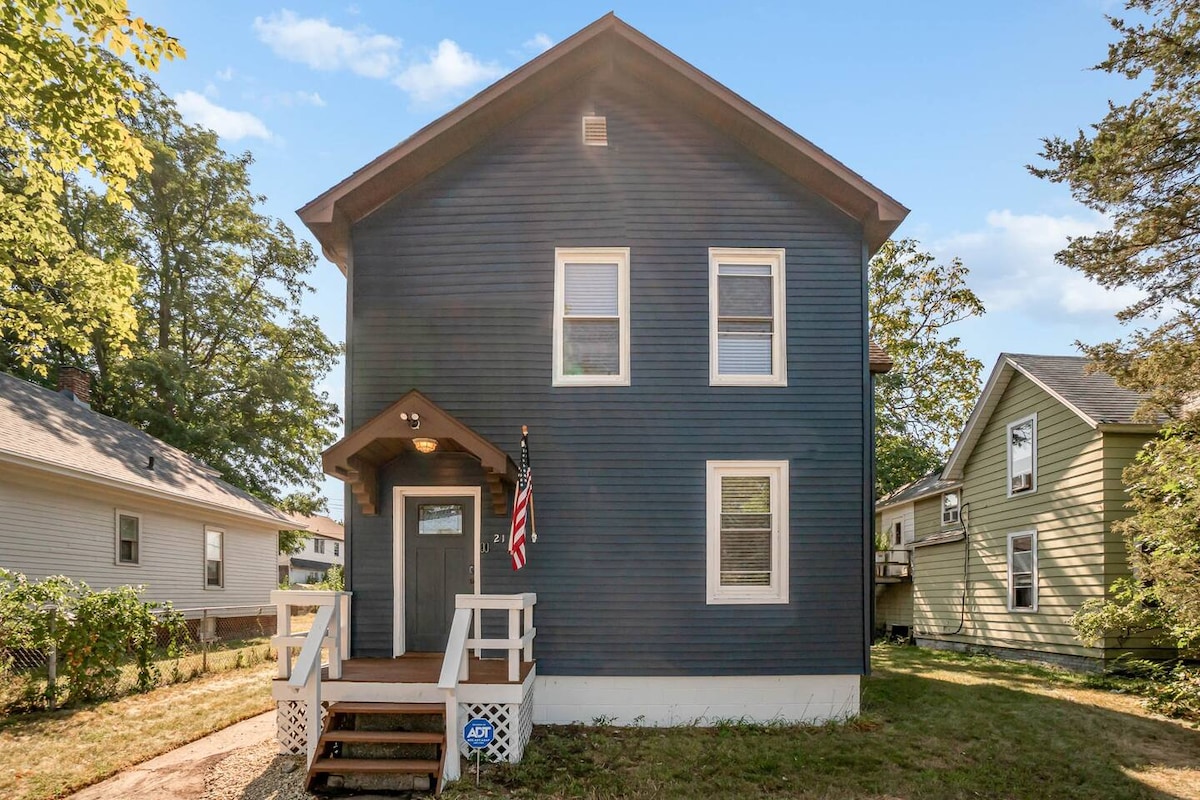 The exterior of a two-story house is shown, featuring dark blue siding and white trim. A welcoming front porch with steps leads to the entrance, which is flanked by a U.S. flag. Trees and neighboring houses provide a quaint setting.