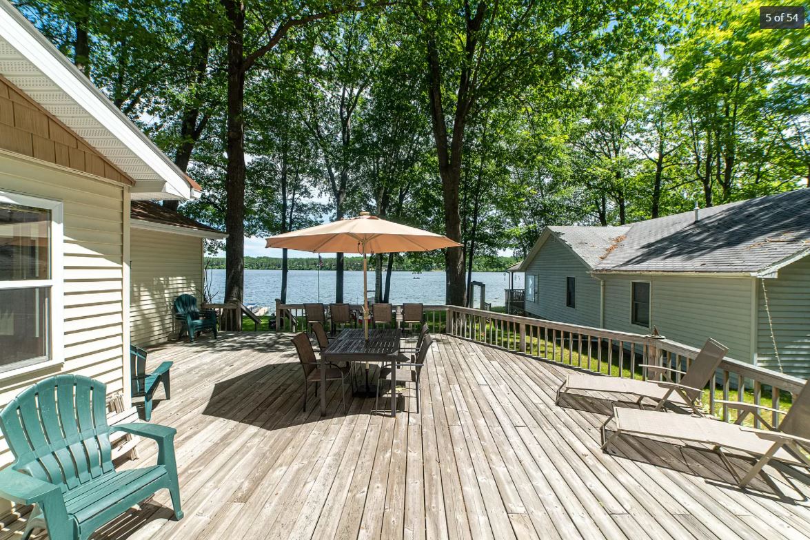 A spacious outdoor deck is surrounded by tall trees, overlooking the lake. Several green Adirondack chairs are positioned around a large table shaded by an umbrella. Sun loungers are placed along the edge, providing a relaxing spot for guests to enjoy the view.