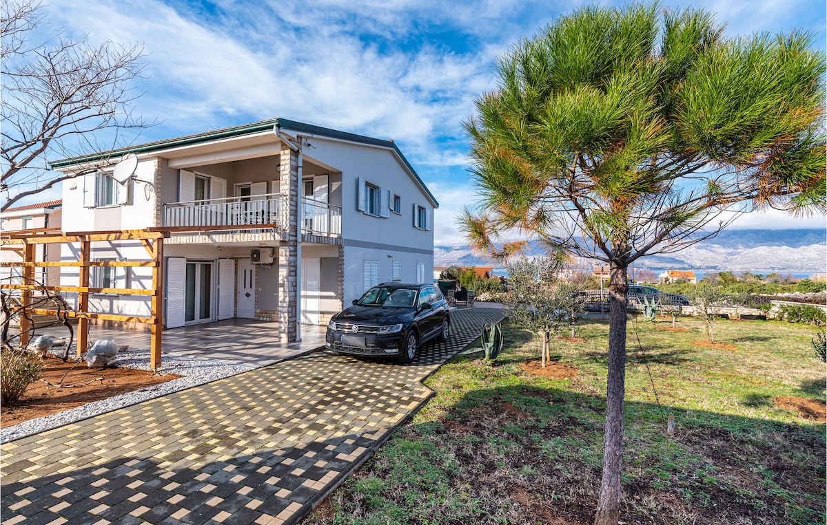 A two-story building is shown, featuring a light-colored exterior and a paved driveway leading to the entrance. A vehicle is parked nearby, while a lone tree stands in the foreground, surrounded by landscaped garden areas with grass and low shrubs.