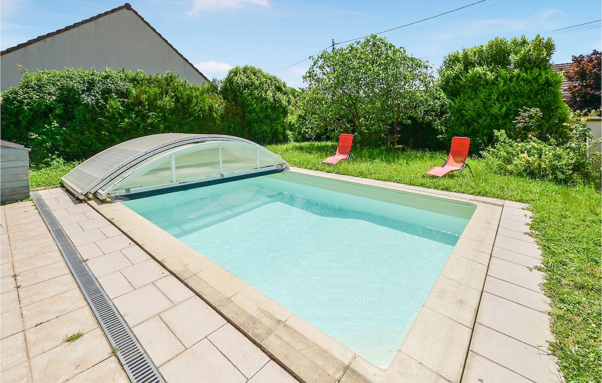 An outdoor private heated pool is situated on a stone patio, surrounded by lush greenery and grass. Two red lounge chairs are positioned nearby, inviting relaxation. A clear cover partially shelters the pool, offering protection while allowing for ample natural light.