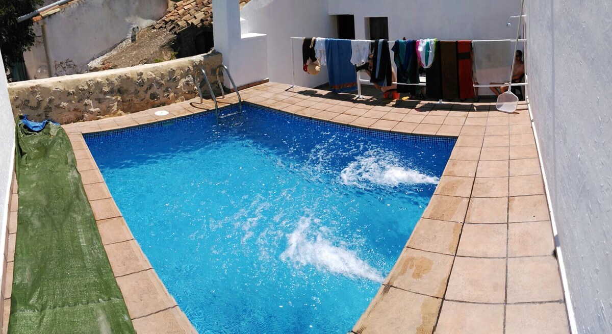 A private swimming pool is visible, surrounded by tiled flooring, with splashes of water indicating activity. Nearby, colorful clothing hangs on a clothesline, providing a practical use of the outdoor space. A grassy area lines one side of the pool, adding a touch of nature.
