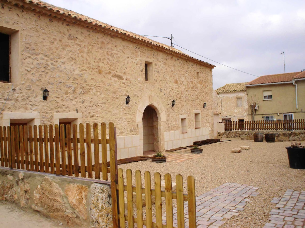 The exterior of the villa features a traditional stone facade with archway entrance. The courtyard is surrounded by a wooden fence and showcases a gravel surface. Nearby, there are several planters and rustic structures, highlighting the inviting, tranquil atmosphere.