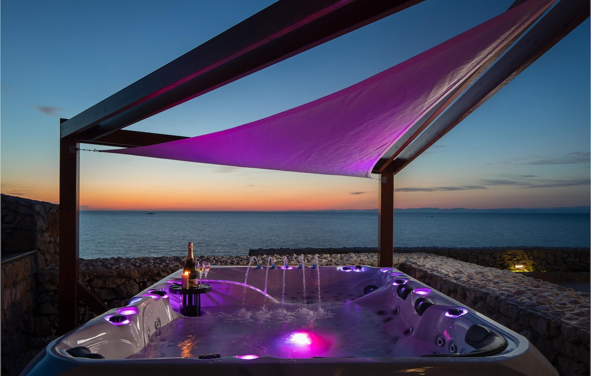 An outdoor hot tub is illuminated with soft purple lights beneath a shaded pergola. The serene sea view extends to the horizon, showcasing a gradient sky as the sun sets. A bottle and glasses are positioned on a nearby table, inviting relaxation.
