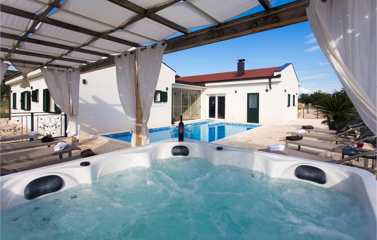 A hot tub is positioned under a shaded pergola, surrounded by stone decking. The background features a pool and a white house with green shutters, providing a serene outdoor space for relaxation. Curtains add privacy, while sun loungers are arranged nearby.