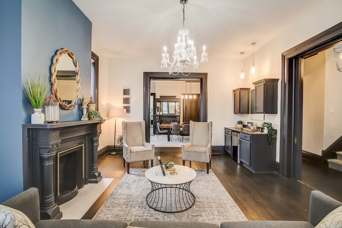A living area featuring a classic fireplace with a decorative mantel serves as a focal point. Light-colored armchairs are arranged around a circular coffee table on a soft rug. An elegant chandelier hangs overhead, and dark wood accents frame the space.