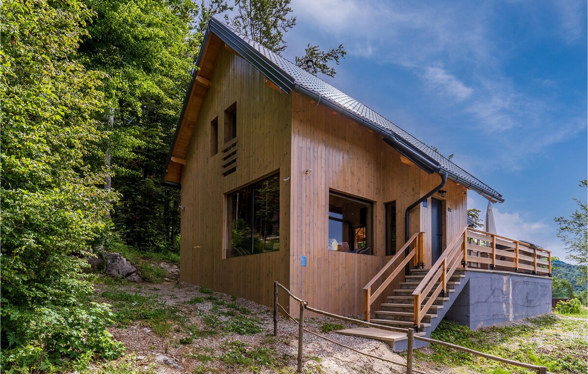 A wooden house is presented amidst lush greenery, featuring a sloped roof and large windows allowing for ample natural light. A set of steps leads to the entrance. The surrounding landscape highlights the tranquil mountain setting, contributing to an inviting natural atmosphere.