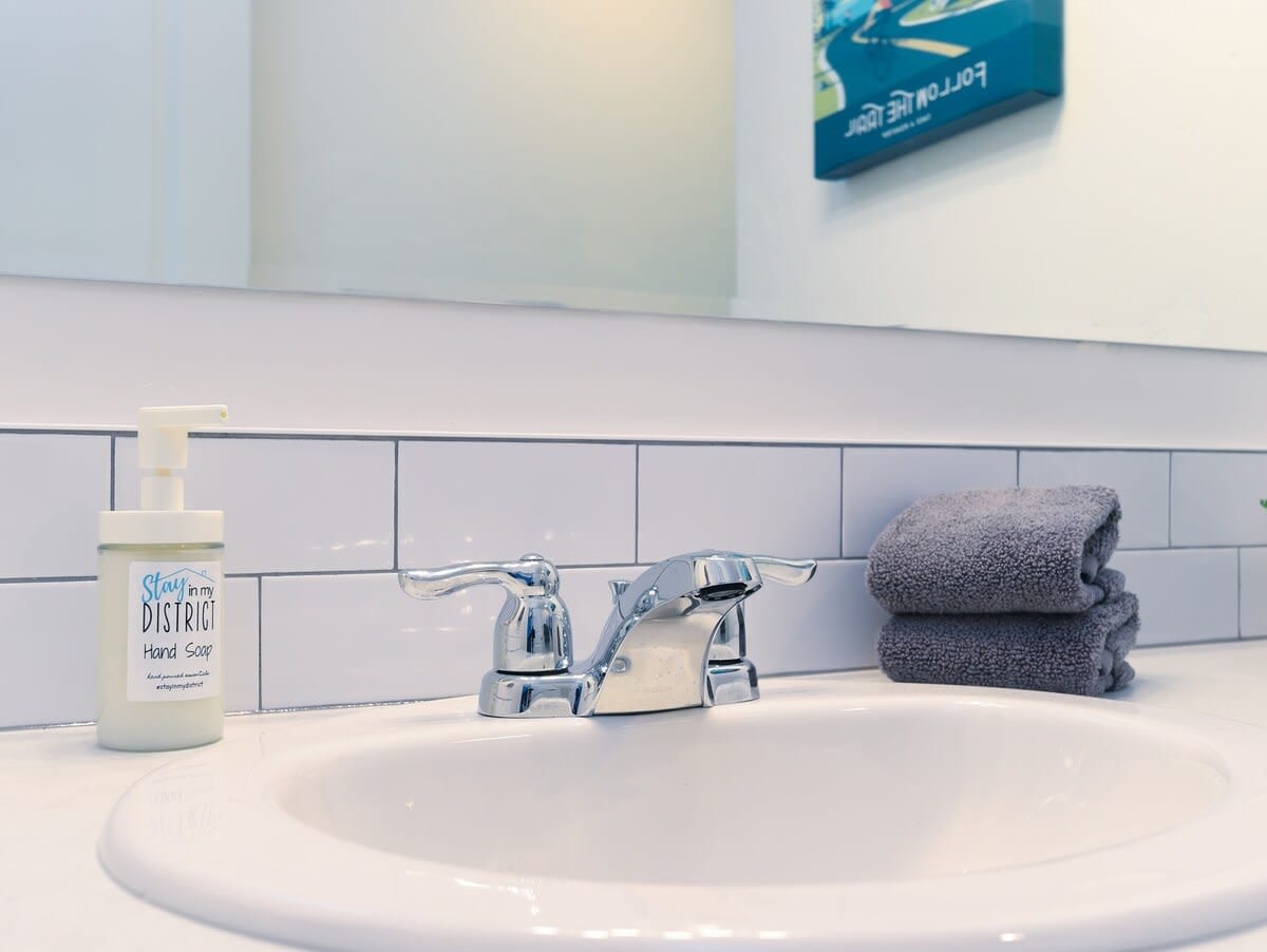 A clean and modern bathroom sink area features a white porcelain basin with a sleek chrome faucet. A bottle of hand soap is positioned next to neatly folded gray towels, providing an organized touch. The simple tile backsplash adds a classic element to the space.