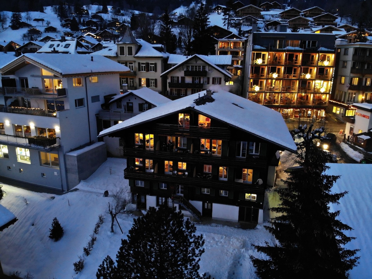 A snow-covered landscape showcases a cluster of buildings in a mountainous area. The architectural styles vary, featuring wooden facades and balconies. Warm lights glow from the windows, creating a cozy contrast against the winter scenery while trees stand tall in the foreground.