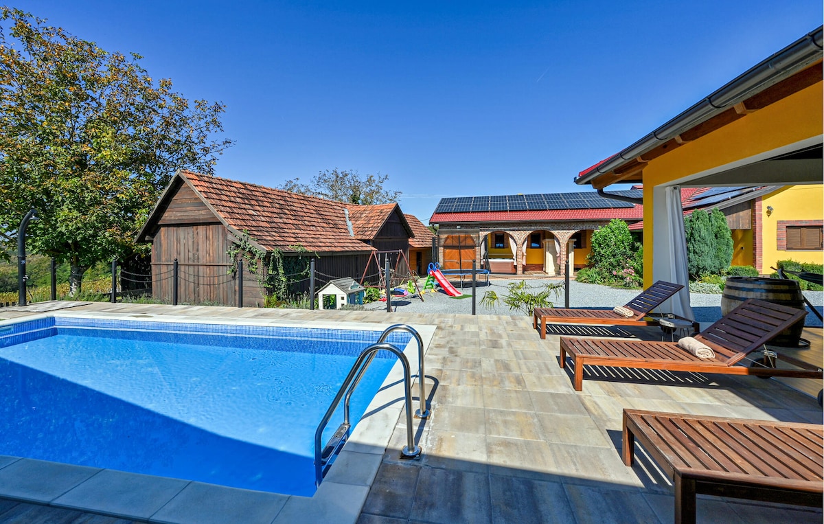 A clear blue sky is seen above a private pool surrounded by stone tiles. Sun loungers are positioned nearby for relaxation. In the background, charming wooden structures are visible, along with colorful playground equipment, providing a family-friendly atmosphere on the spacious property.
