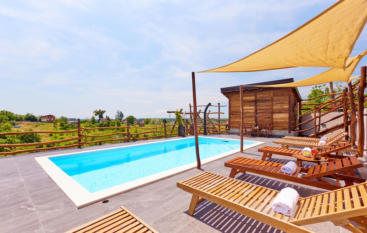 A sunlit outdoor area features a refreshing swimming pool surrounded by wooden loungers. Sun shades cast gentle shadows over the space. The backdrop showcases rolling hills and green landscapes, enhancing the serene environment for relaxation.