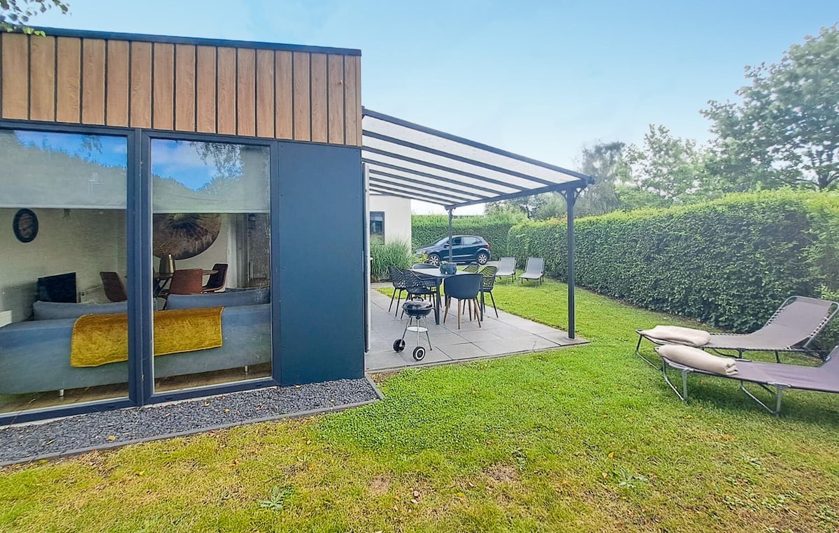 The exterior of the cottage reveals a spacious patio area shaded by a pergola. Outdoor seating is arranged around a large table, accompanied by lounge chairs. A grassy area is visible, framed by lush greenery, providing a welcoming space for relaxation.