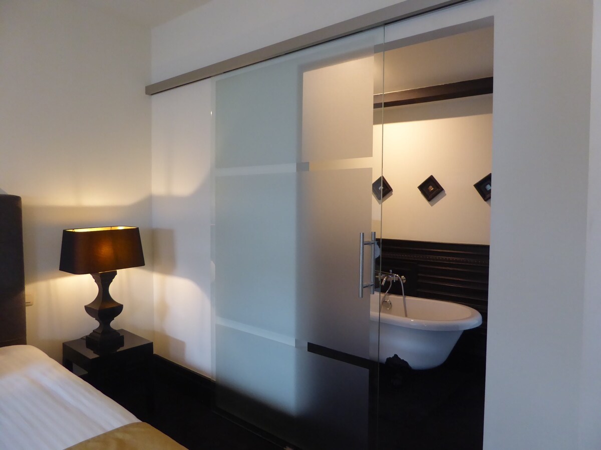 A spacious room is complemented by a sleek glass door leading to a stylish bathroom. Soft light from a bedside lamp casts a warm glow, while a classic freestanding bathtub is visible through the frosted glass, enhancing the room's modern aesthetic.