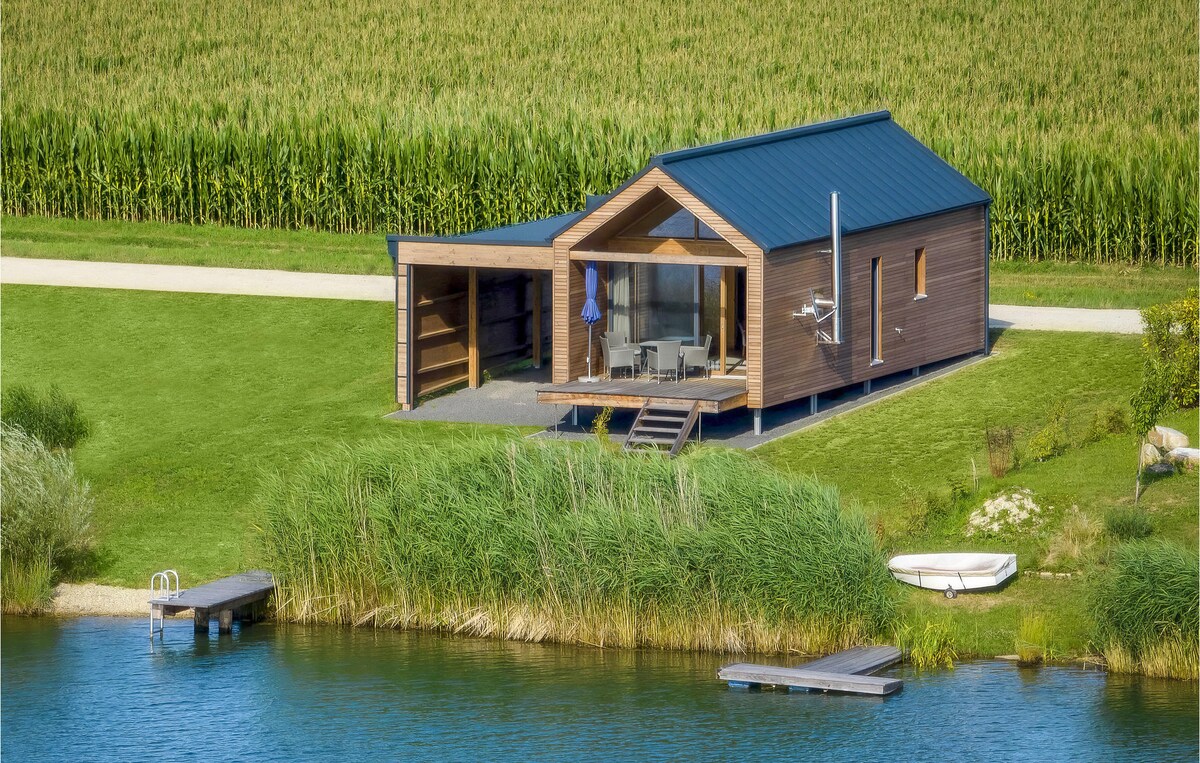 The vacation home is surrounded by lush greenery, positioned near a tranquil lake. A covered terrace is visible, furnished with seating and an umbrella. A wooden dock extends from the lakeside, leading to a small boat, while the natural landscape provides a serene backdrop.