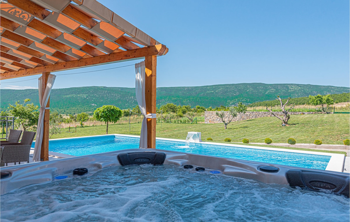 A hot tub is situated beneath a wooden covered area, offering views of a landscaped yard and distant hills. Nearby, a private swimming pool reflects the clear blue sky, while a serene atmosphere is enhanced by the surrounding greenery.