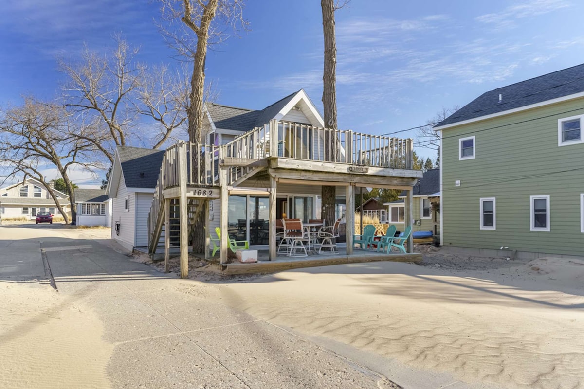 The beach cottage is showcased with a covered upper deck and a spacious outdoor area. The two-story structure is flanked by sandy surroundings, featuring large glass windows that provide views of the nearby beach. Colorful outdoor seating is visible on the deck.