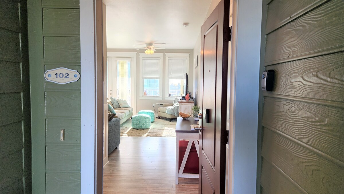 A view from the entryway showcases the bright living area, featuring a comfortable sofa and accent chairs. Large windows allow natural light to fill the space, and tasteful decor elements add a welcoming touch. The flooring is a warm wood finish.