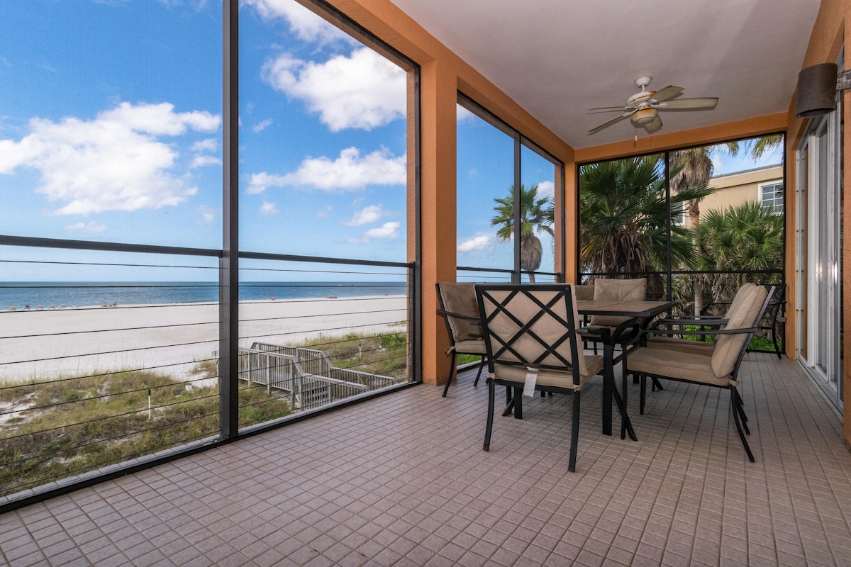 A spacious screened lanai is shown, featuring a dining table surrounded by six chairs. The backdrop includes expansive views of the beach and sea, with palm trees framing the scene. Soft cloud patterns are visible in the bright blue sky.