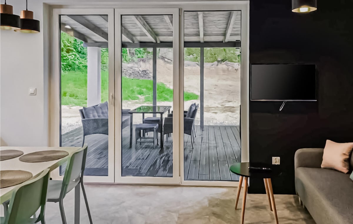 A modern living area is featured, with large sliding glass doors opening to a covered terrace. Outdoor seating and a dining table are visible through the doors, framed by greenery. A simple dining table is set nearby, complemented by a small side table and a sofa.