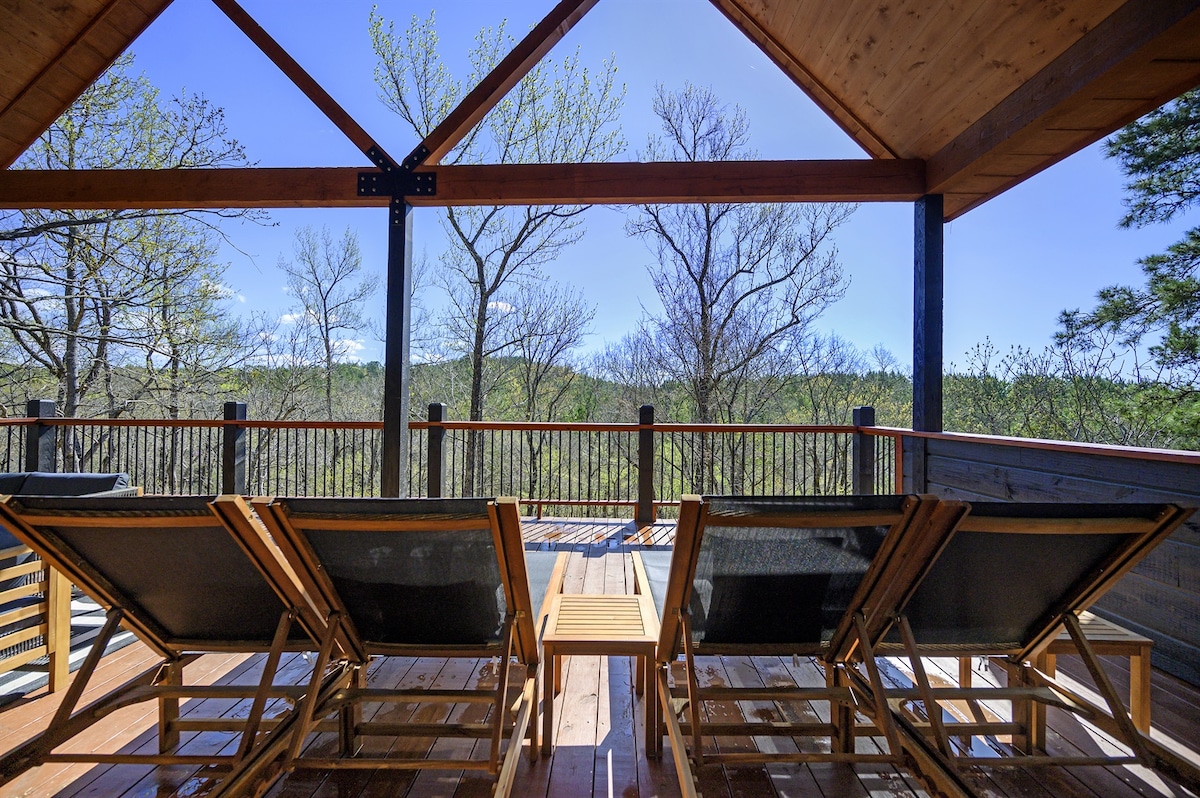 A covered outdoor deck features two wooden lounge chairs positioned toward a scenic view of trees and hills. A small table is placed between the chairs, highlighting the connection to nature in a serene setting.