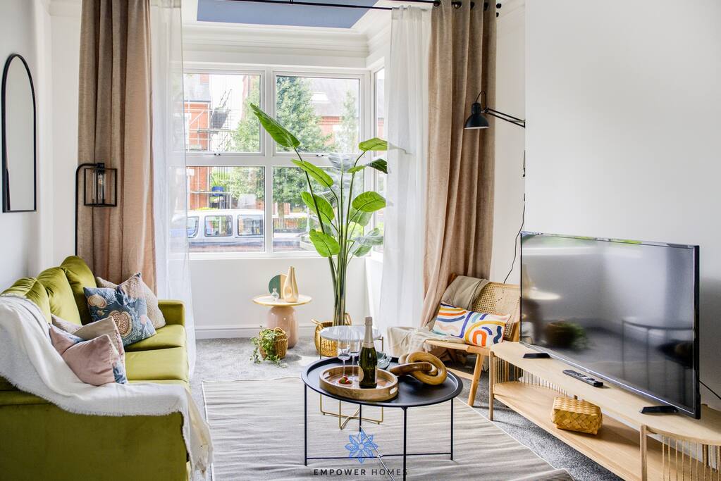 The open lounge area features large windows allowing natural light to fill the space. A green sofa and beige armchair are arranged around a circular coffee table. A tall plant adds a touch of nature, while a flat-screen TV is positioned on a light wood media console.