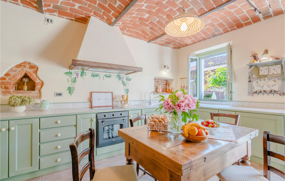 A charming kitchen displays green cabinetry with a rustic wooden table at its center. Fresh fruits and flowers add a touch of nature, while a vintage stove occupies a prominent corner. Exposed brick ceilings and a window provide warmth and light to the space.