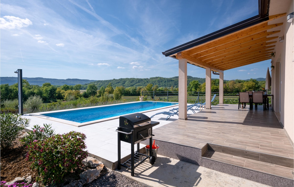 The covered terrace features a barbecue grill, with a view of the outdoor pool and surrounding greenery. Sun loungers are positioned beside the pool, and the landscape includes rolling hills and trees under a clear blue sky.