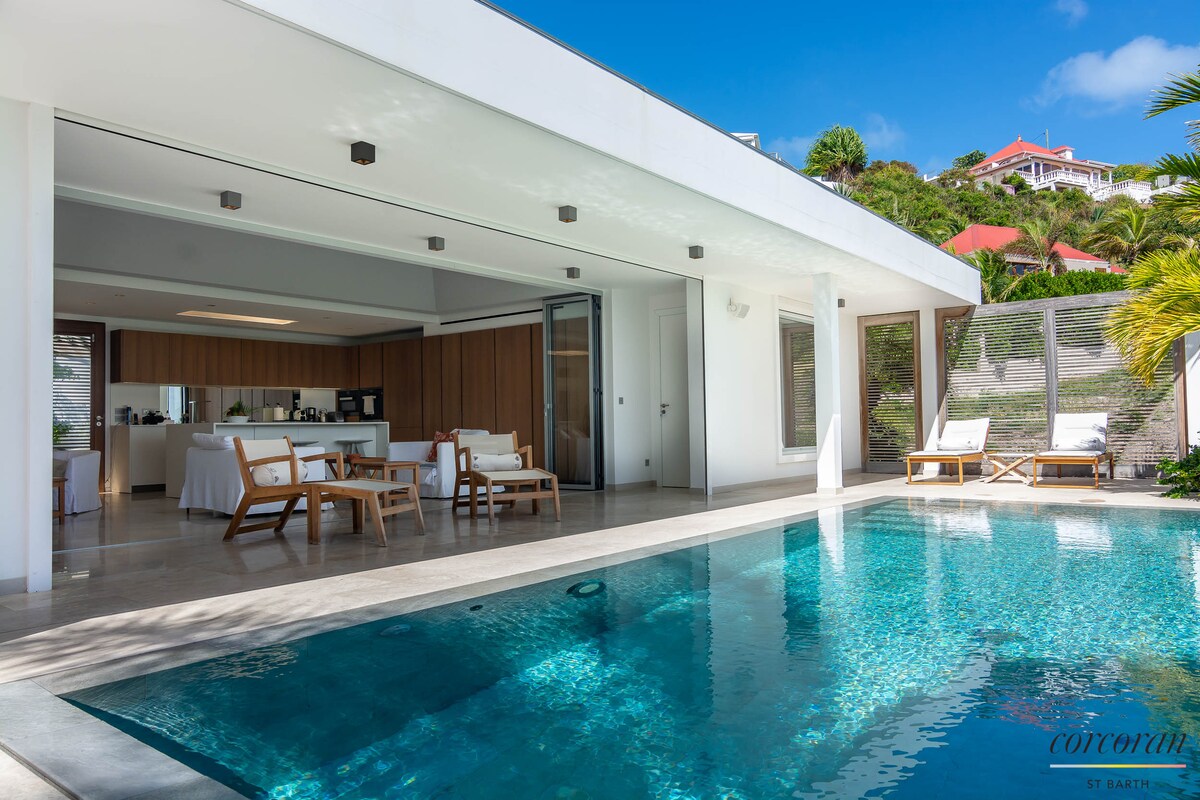 An open living area is visible with a sleek design, featuring a modern kitchen and a spacious heated pool that reflects the clear blue sky. Comfortable loungers surround the pool, nestled against the natural greenery of the surrounding hillside.