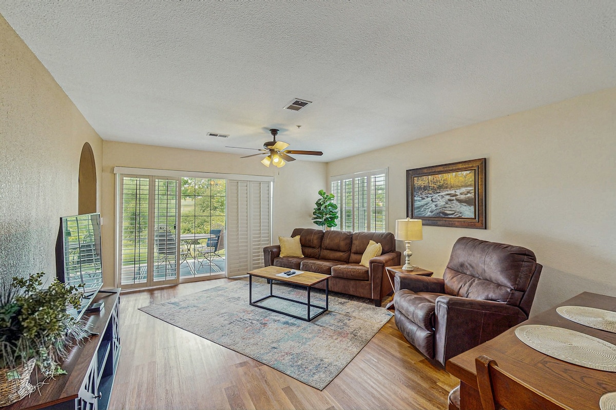 A sunlit living area features comfortable seating, including a recliner and a sofa. A coffee table is centered on a decorative area rug. Light streams through windows with blinds, while a patio view showcases the lush outdoors. A painting adds an artistic touch on the wall.