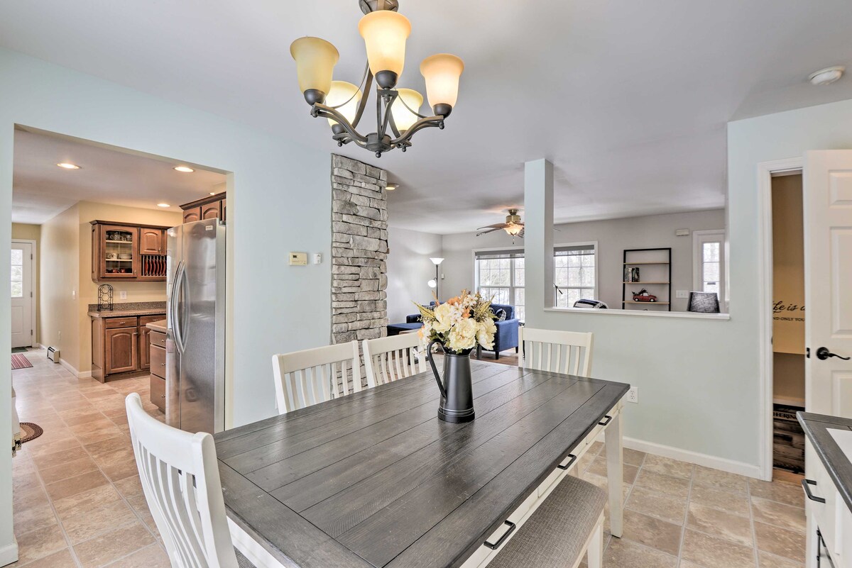 A bright dining area is shown, featuring a large rectangular table surrounded by six chairs. A chandelier with multiple bulbs hangs above, and a vase with fresh flowers is centered on the table. The adjacent kitchen is visible through an open doorway.