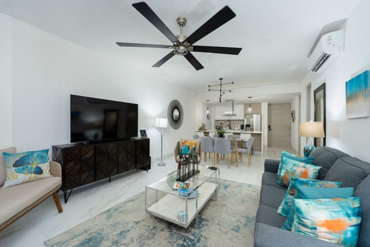 The living area features a light-filled, modern design with a large flat-screen TV mounted on a stylish cabinet. Comfortable seating is arranged around a coffee table, adorned with decorative items. A dining space is visible in the background, showcasing a well-lit open layout.