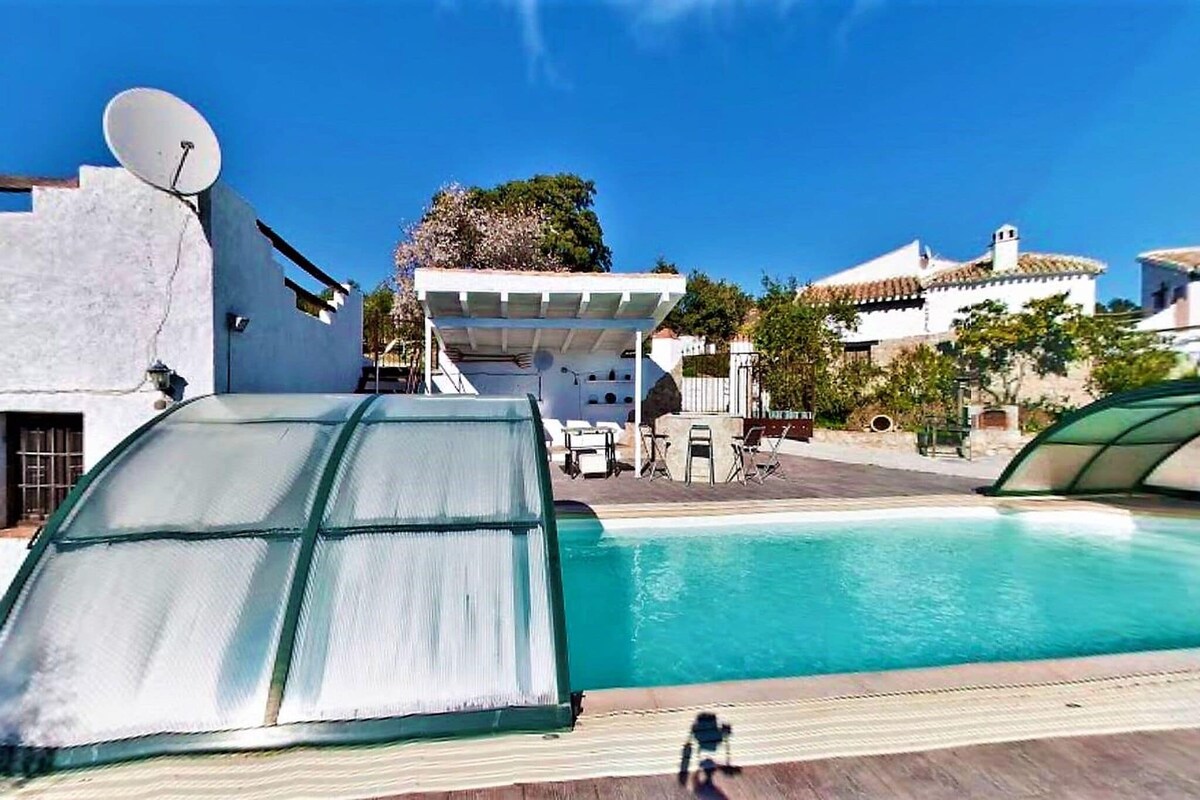 The outdoor swimming pool is enclosed with a retractable cover, surrounded by wooden decking. A shaded seating area with lounge furniture is visible in the background, offering a relaxing space under the clear blue sky. Green trees and plants provide a natural ambiance.