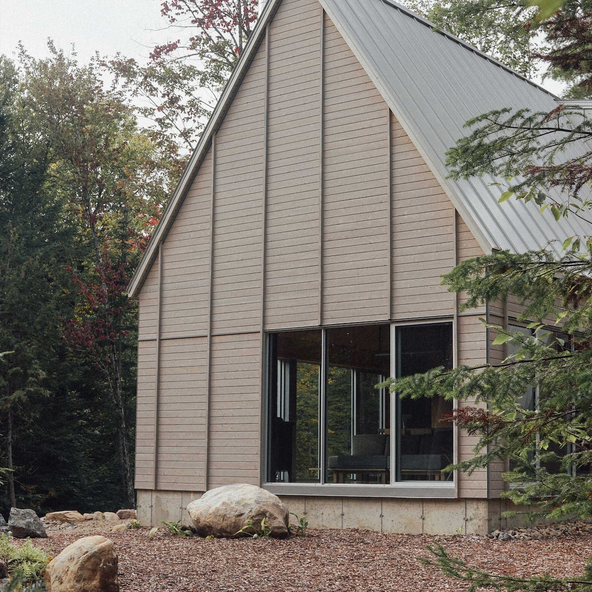 The exterior of the chalet showcases a modern design with wooden paneling and a sloped roof. Large windows offer a glimpse of the interior, surrounded by natural landscaping featuring rocks and trees that complement the wooded environment.