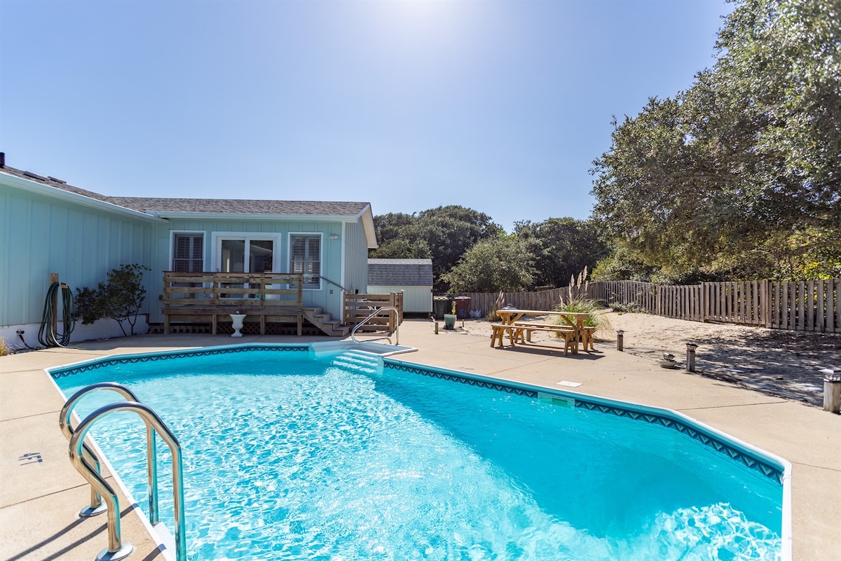 A private, heated saltwater pool is surrounded by a spacious patio with sunlight reflecting off the water. The inviting outdoor space includes seating arrangements and a backdrop of lush greenery and sand, offering a tranquil retreat just steps from the beach cottage.