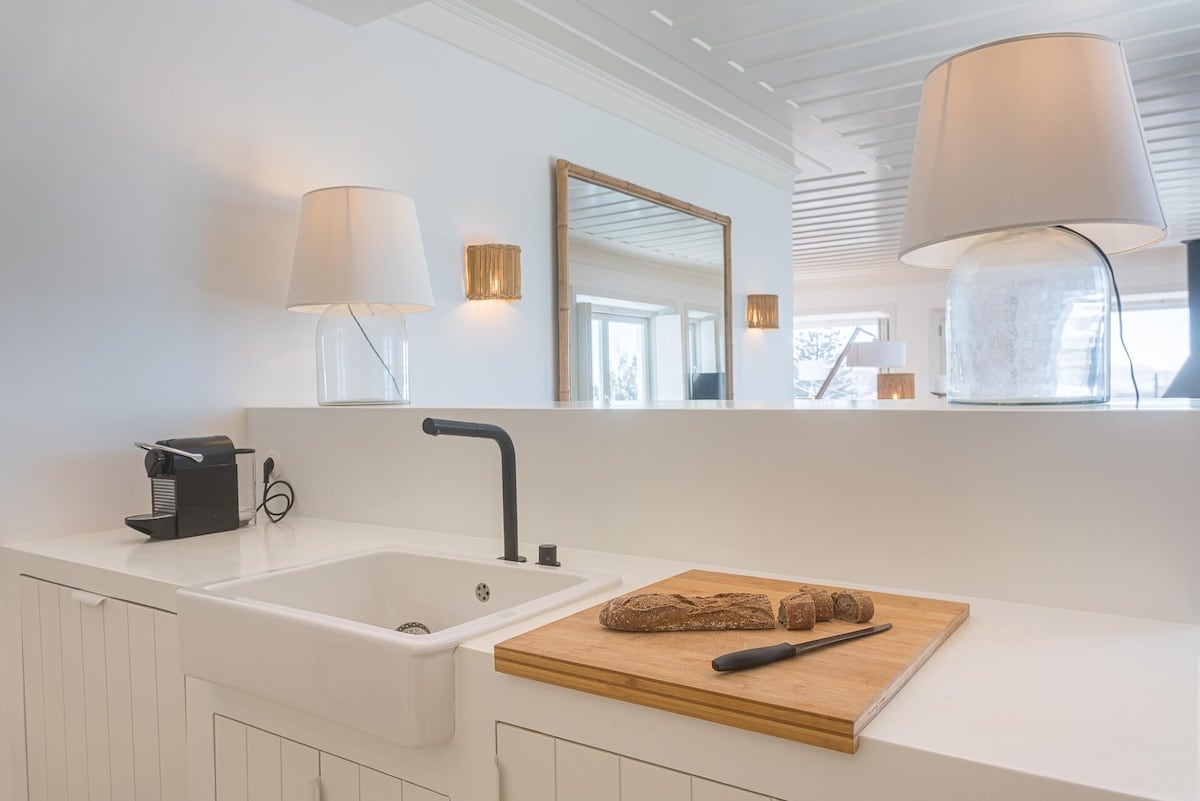 A modern kitchen counter is shown with sleek cabinetry and a white sink. A cutting board with sliced bread and a knife rests on the counter. Two elegant lamps illuminate the space, with a mirror reflecting light from a window nearby.