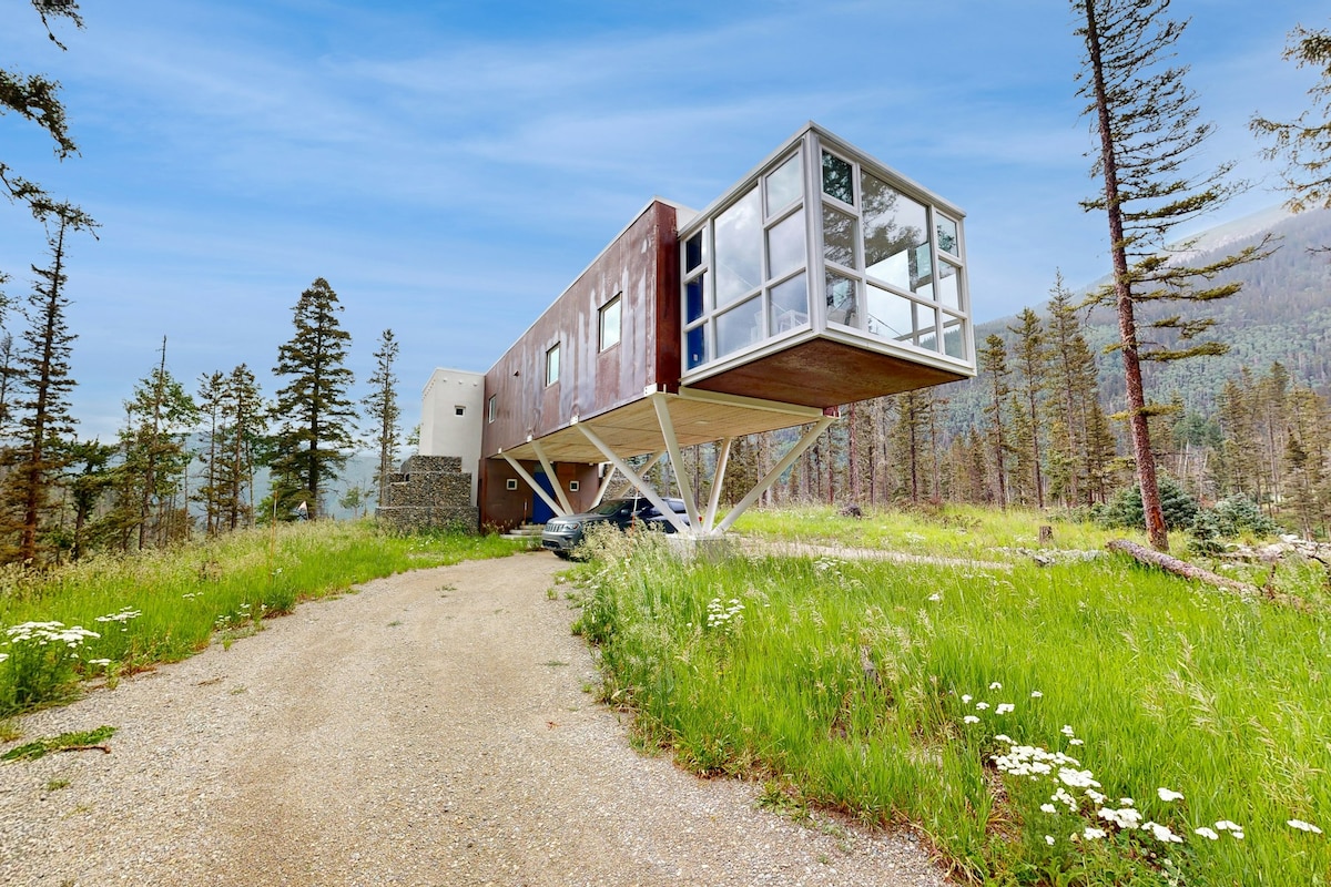 The modern two-story home features a unique cantilevered design, elevated on metal supports. Large glass windows dominate one side, offering views of the surrounding greenery. Natural landscaping includes wildflowers and trees, creating a serene environment. A gravel path leads up to the entrance.