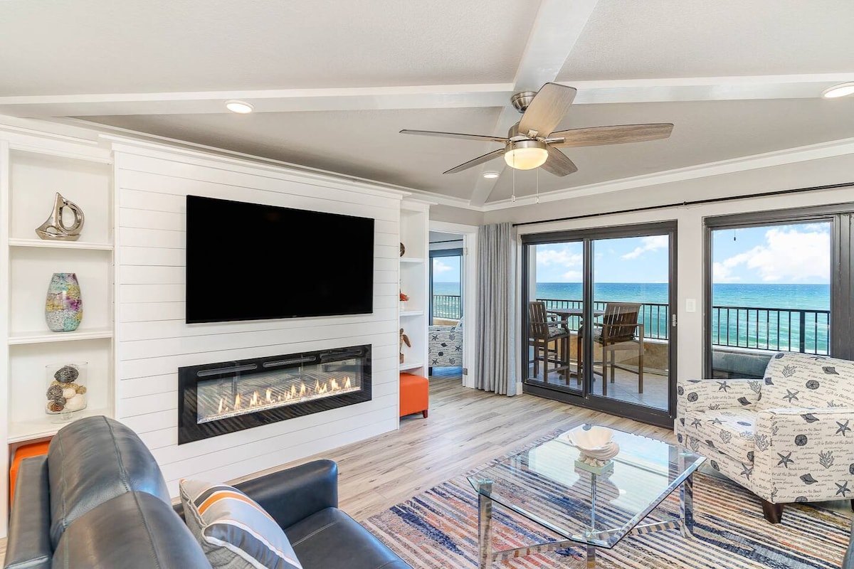 A modern living area features a sleek fireplace and a large wall-mounted flat-screen TV. A comfortable dark sofa and a patterned armchair provide seating. Large sliding glass doors open to a balcony with ocean views, allowing natural light to brighten the space.