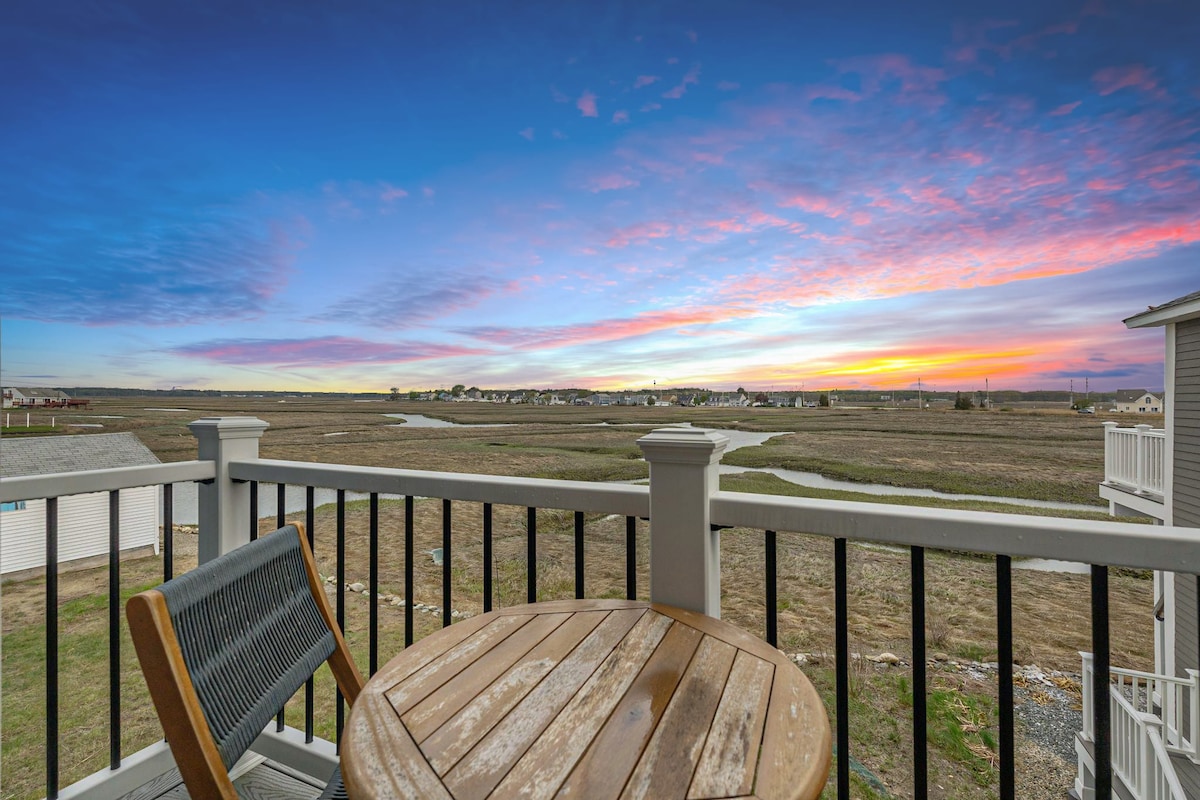 A small balcony features a two-seater wooden table with a circular top. This outdoor area provides unobstructed views of the expansive salt marsh, where the sky transitions into vibrant hues of orange, pink, and blue during sunset.
