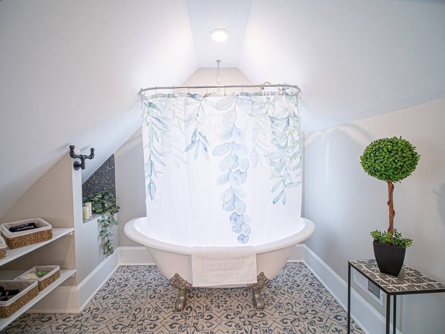 A stylish bathroom features a clawfoot tub elegantly positioned beneath a sloped ceiling. A floral shower curtain provides a soft visual touch, while decorative green foliage adds a natural element. Shelving units hold assorted baskets, enhancing the organized space.