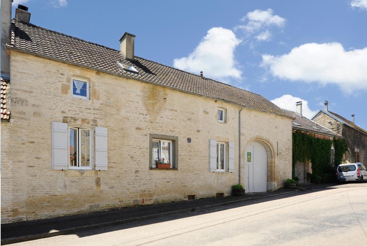 A charming stone building is displayed, featuring a pitched roof and white shutters. A wooden door is set within an arched entryway, while a window is adorned with small decorative elements. The exterior is complemented by greenery climbing the walls, with a quiet street visible in front.