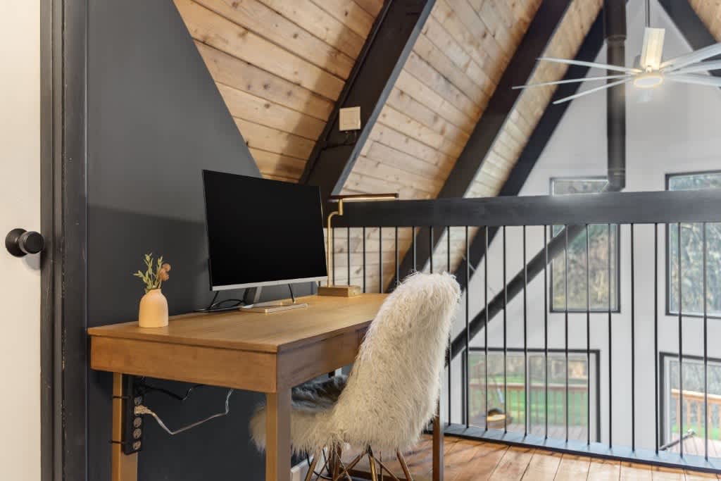 A workspace is showcased with a wooden desk paired with a chair featuring a soft white cushion. A computer monitor is positioned on the desk, and large windows offer views of the outdoor landscape. The sloped ceiling is accented by wooden beams.