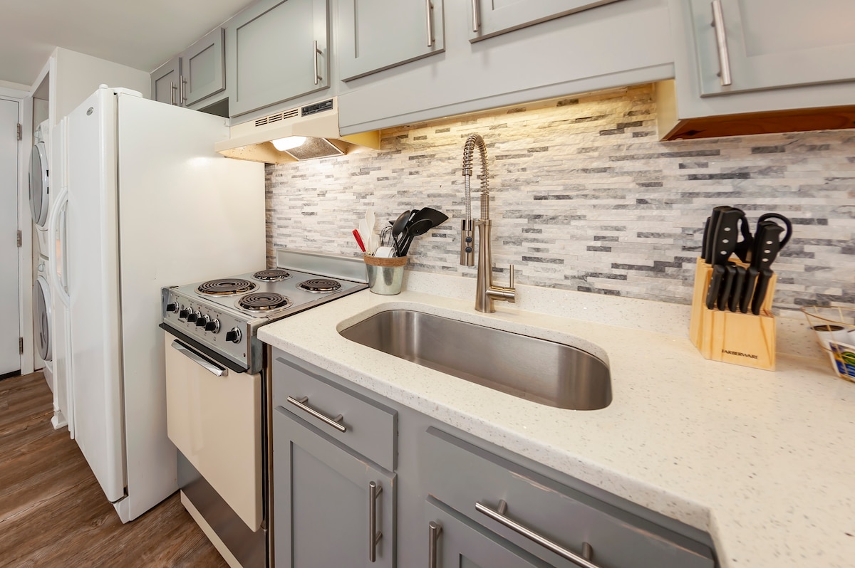 The kitchen is equipped with modern appliances, including a stove and refrigerator. A sleek countertop complements the cabinetry, while a faucet and sink are positioned near essential cooking tools in a utensil holder, creating an efficient workspace.