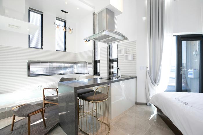 A modern interior is featured with a sleek kitchen island, gold-accented stools, and ample natural light from the large windows. A cozy bed is positioned against the wall, and a sliding door leads to a private balcony.